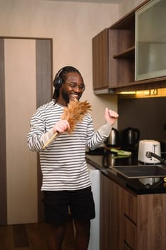 Carefree young African man listening to music in headphones and singing while doing housework.