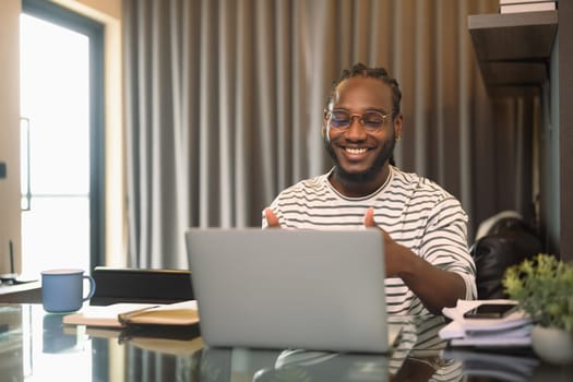 Positive African man looking at laptop talking to online video call distant speaking with friends.