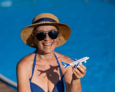 An elderly woman in a hat and sunglasses holds a model airplane while sitting by the pool. Retirement vacation concept