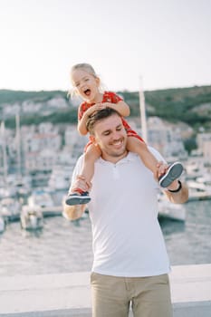 Dad with a laughing little girl on his shoulders stands at the boardwalk fence with his eyes closed. High quality photo