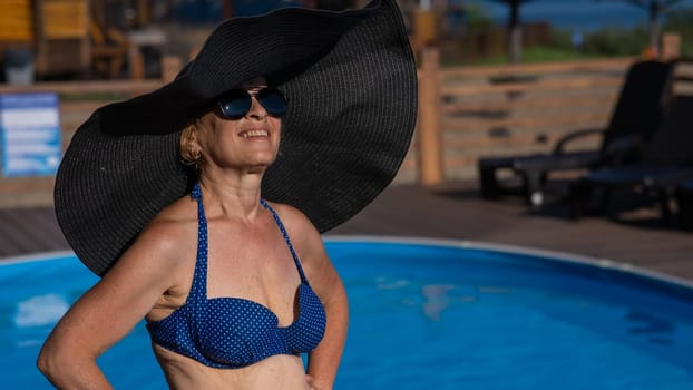 An elderly woman in a huge straw hat and sunglasses is sunbathing by the pool. Retiree on vacation