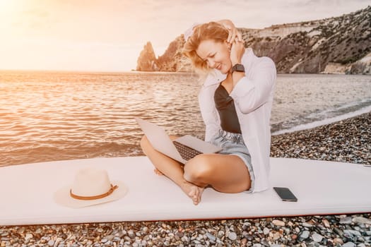 Successful business woman in yellow hat working on laptop by the sea. Pretty lady typing on computer at summer day outdoors. Freelance, travel and holidays concept.