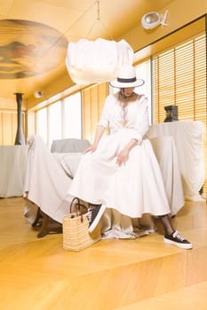 Elegant fashionable woman wearing summer white dress, straw hat, posing in stylish boho interior. Copy, empty space for text