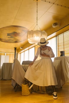 Elegant fashionable woman wearing summer white dress, straw hat, posing in stylish boho interior. Copy, empty space for text