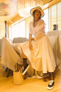 Elegant fashionable woman wearing summer white dress, straw hat, posing in stylish boho interior. Copy, empty space for text