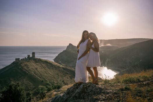 Two young girls are standing on a hillside, one of them wearing a white dress. The sun is shining brightly, creating a warm and inviting atmosphere. The girls seem to be enjoying their time together