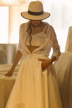 Elegant fashionable woman wearing summer white dress, straw hat, posing in stylish boho interior. Copy, empty space for text