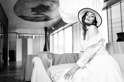 Elegant fashionable woman wearing summer white dress, straw hat, posing in stylish boho interior. Copy, empty space for text