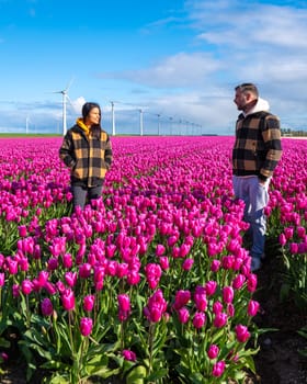 In a vibrant field of purple tulips in the Netherlands, two people stand enchanted by the beauty of Spring, surrounded by windmill turbines. couple of men and women in a flower field