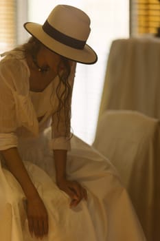 Elegant fashionable woman wearing summer white dress, straw hat, posing in stylish boho interior. Copy, empty space for text