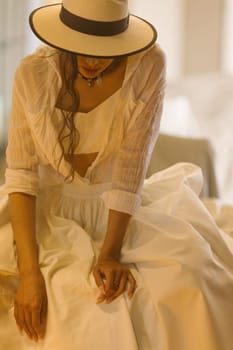 Elegant fashionable woman wearing summer white dress, straw hat, posing in stylish boho interior. Copy, empty space for text