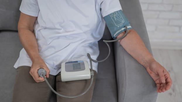 Mature Caucasian woman measuring blood pressure while sitting on sofa at home