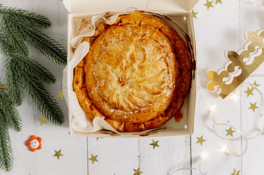 One royal galette in an open box with a golden crown, fir branches, a burning garland, a surprise gift and scattered confetti stars lies on a white wooden table, flat lay close-up.