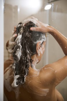Close-up Rear view of pretty woman shampooing hair, showering with moisturizing foam for skin care and body freshness in a douche cabin. Home spa. Beauty, daily hygiene and purity concept