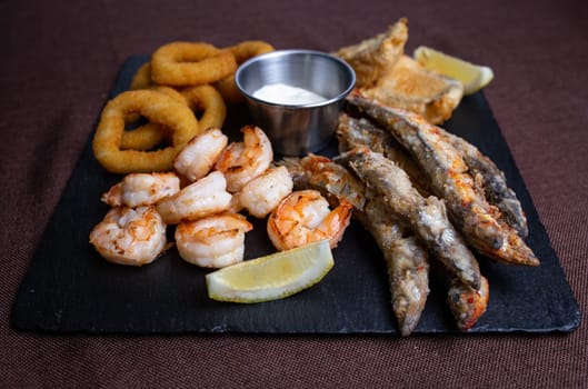 A delicious and healthy meal of fried seafood, including shrimp, fish, and calamari, served with lemon wedges on a black slate plate.