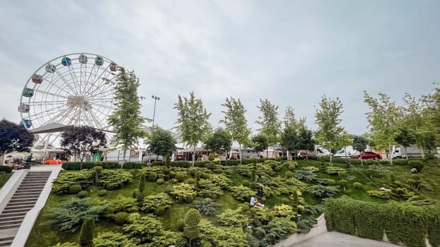 At the amusement park, a Ferris wheel stands tall amidst green trees under a cloudy sky, creating a picturesque scene.