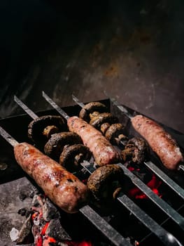Four skewers of sausages and mushrooms over hot coals on a grill grate with smoke rising in the background
