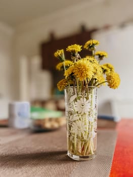 The dandelions are in full bloom and the vase is sitting on a brown table. The background is blurry and light colored.