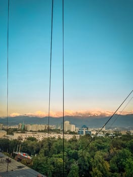 A stunning cityscape with snowy mountains, tall buildings, and green trees under a clear blue sky with a hint of sunset.