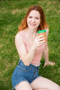 A beautiful young woman with braces on her teeth drinks from a cardboard cup while sitting on the grass. Vertical photo