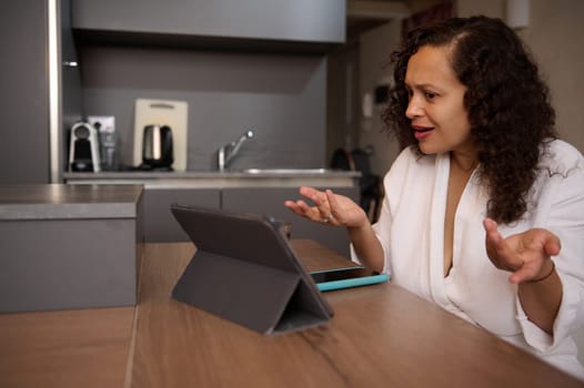 Young multi ethic woman in white bathrobe, discussing with friends or relatives over video call, sitting at table at home. Video conference on digital tablet. Online communication. People and internet