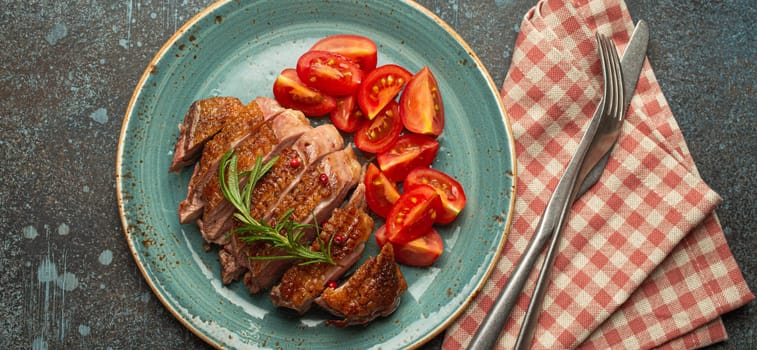 Delicious roasted sliced duck breast fillet with golden crispy skin, with pepper and rosemary, top view on ceramic blue plate served with cherry tomatoes salad, rustic concrete rustic background.