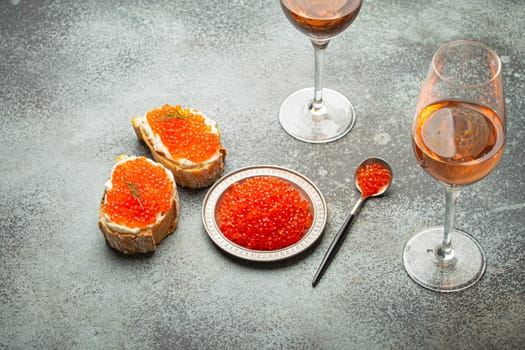 Small metal plate with red salmon caviar, two caviar toasts canape, two glasses of champagne top view on grey concrete background, festive luxury delicacy and appetizer.