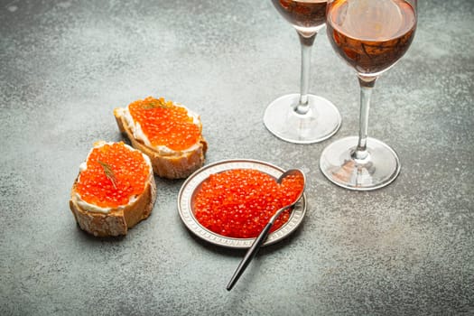 Small metal plate with red salmon caviar, two caviar toasts canape, two glasses of champagne top view on grey concrete background, festive luxury delicacy and appetizer.