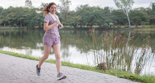 A woman is running in a park near a lake. She is wearing a purple shirt and shorts