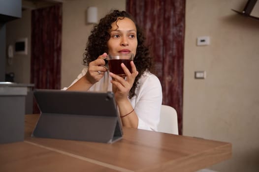 Multi ethnic beautiful woman in white bathrobe, holding cup of tea, dreamily looking aside, sitting t kitchen table in front of digital table in modern home interior. People. Communication. Internet.