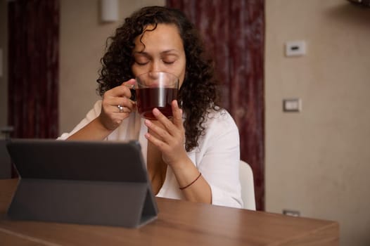 Beautiful multi ethnic middle aged woman in white bathrobe, drinking hot tea, sitting at digital tablet at home interior. People. Lifestyle