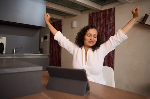 Charming young woman smiling stretching up after waking up in the morning, sitting at table with a digital tablet. People. Morning routine. Lifestyle. Online communication and business. Remote job