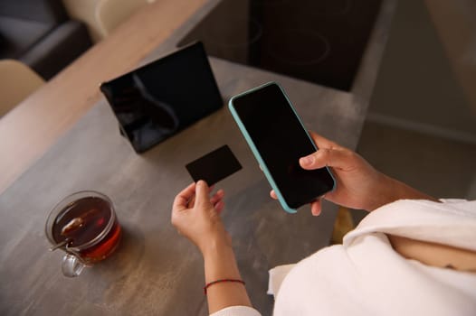 Close-up woman holds credit card, using smartphone with black mockup touch screen, online shopping. Businesswoman freelancer receives payment, checks balance on mobile app, booking tickets on internet