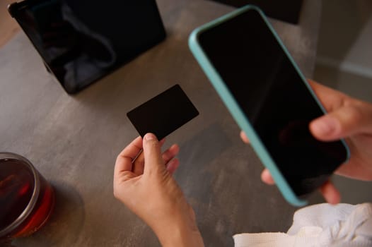 Close-up woman's hands hold a black mockup credit card with copy ad space for text, and using smart phone for online shopping, receiving or making payments via internet mobile banking application