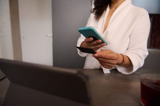 Close-up view of a woman in bathrobe at home, holding blank black credit card, using mobile phone, transferring money via internet banking, making payment, using mobile app. Copy advertising space