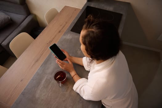 Overhead view of young adult woman holding a smartphone with blank black mockup touch screen, making online payment, shopping, paying bills, sitting at kitchen table in the home office. Copy ad space