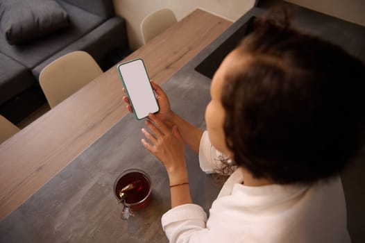 Rear top view of young adult woman holding a smartphone with blank white mockup touch screen, making online payment, shopping, paying bills, sitting at kitchen table in the home office. Copy ad space