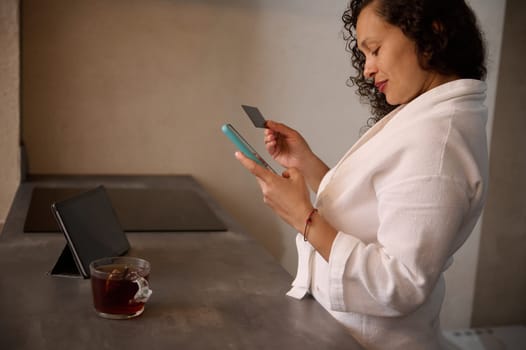 Side portrait of a young y woman in white bathrobe holding black credit card and smartphone, shopping online, making payments on internet via mobile banking, standing t kitchen counter in the morning