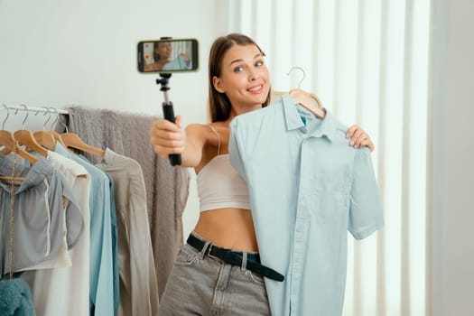 Young woman making fashion video content for social media. Blogger smiles to camera using selfie stick and light ring while making persuasive online clothing sell to audience or followers. Unveiling