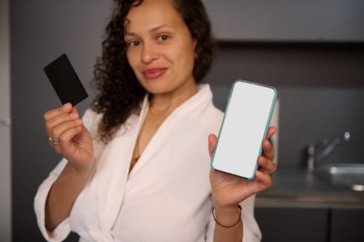 Young woman holds and shows at camera blank black credit card and smartphone with white empty mockup digital screen, makes online payments, transferring money via internet mobile banking or mobile app