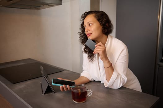 Young curly haired brunette pretty woman in white bathrobe at home, making online payments via internet mobile banking, sitting at table with smart phone and credit card in her hands