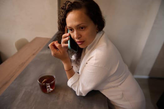 Young multi ethnic woman looking aside, talking on smart mobile phone while taking breakfast at home, standing at kitchen counter in at home, dressed in white bathrobe. Close-up side portrait