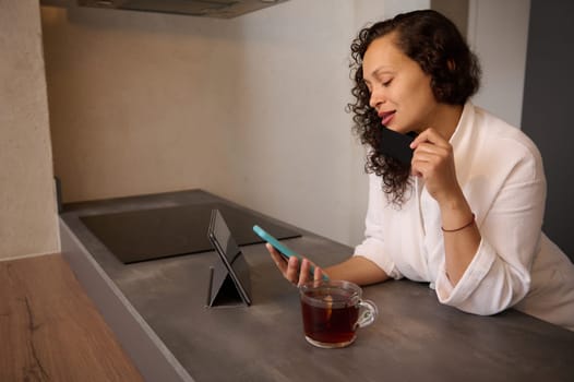Beautiful young woman checking messages on digital tablet, sitting over a cup of hot herbal healing tea. Multi ethnic curly haired brunette in white bathrobe reading news on smartphone, browsing web