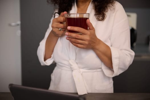 Cropped view of hands of woman in white bathrobe, holding a cup of hot tea