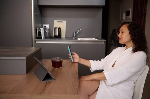 Relaxed young multi ethnic woman in white bathrobe, using mobile phone at home, checking social media content, new mobile application, booking, ordering food, scrolling news feed, online shopping