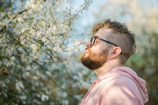 Portrait of curly millennial man inhales the fragrance of spring flowers of blooming jasmine or cherry tree. Spring time