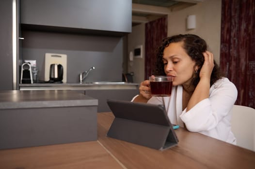 Beautiful young woman in white bathrobe, sitting at kitchen table and drinking tea, watching movie or webinar on digital tablet