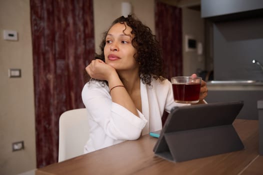 Relaxed young beautiful woman in white bathrobe, holding cup of tea, dreamily looking aside, sitting t kitchen table in front of digital table in modern home interior. People. Communication. Internet.