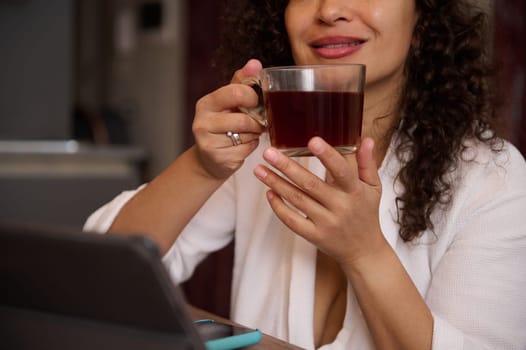 Details on hands of a blurred smiling pretty woman holding a cup of hot tea drink, sitting at table with digital tablet in modern home interior. People. Leisure. Lifestyle. Domestic life
