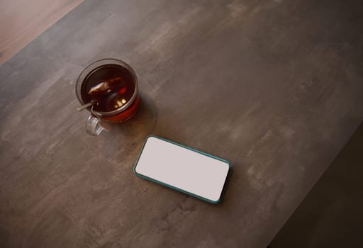 Directly above smartphone white blank digital touch screen mockup copy space for advertising or mobile apps, application, next to a cup of tea on a table. Digital technology. Electronics. Lifestyle